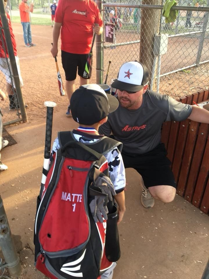 Baseball coach in gray shirt kneels by a young player with red-and-black gear at the dugout fence