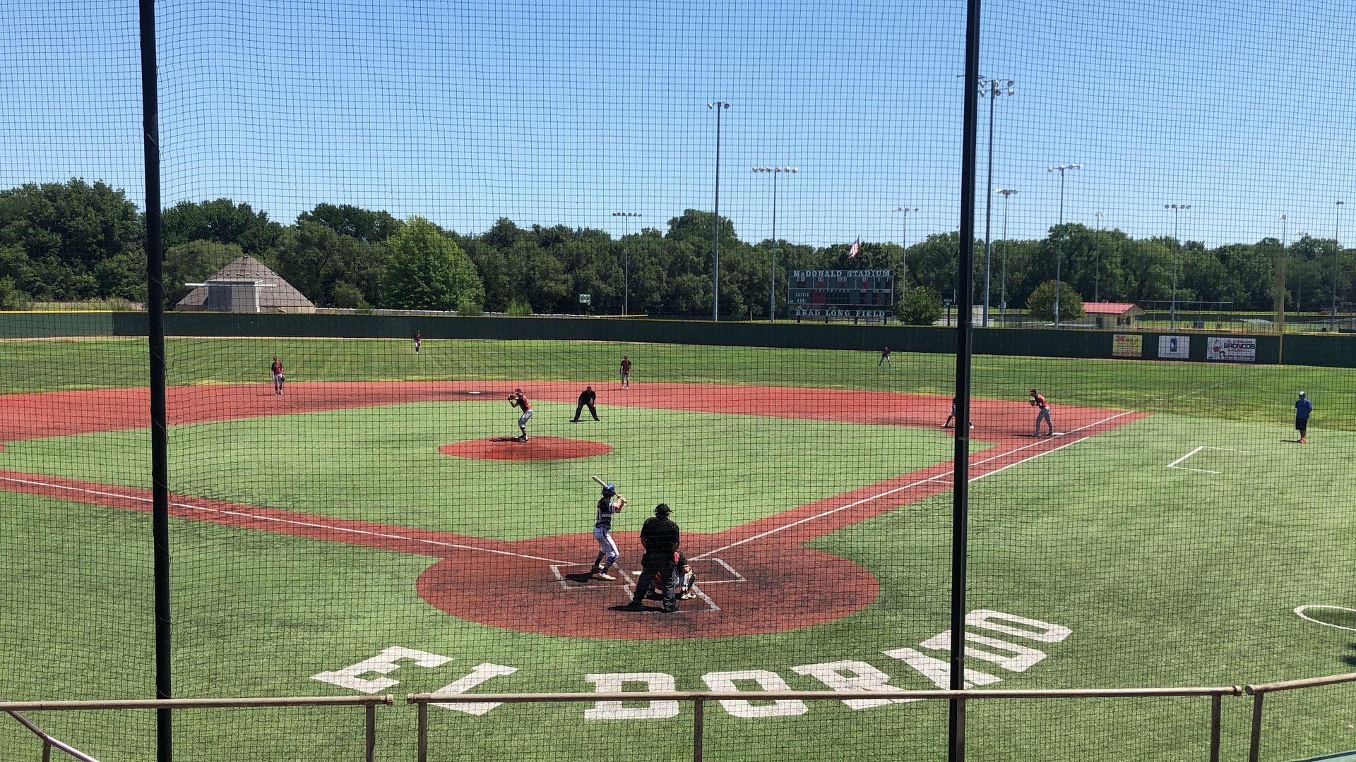 Youth baseball game on a sunny field, viewed from behind home plate with pitcher, batter, and catcher visible.