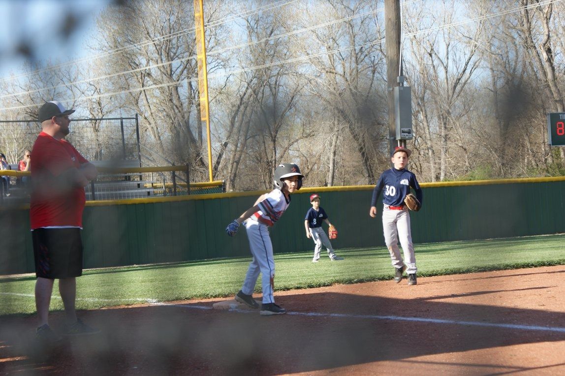 Youth baseball players on a field, with one batter at home plate and fielders in the outfield.