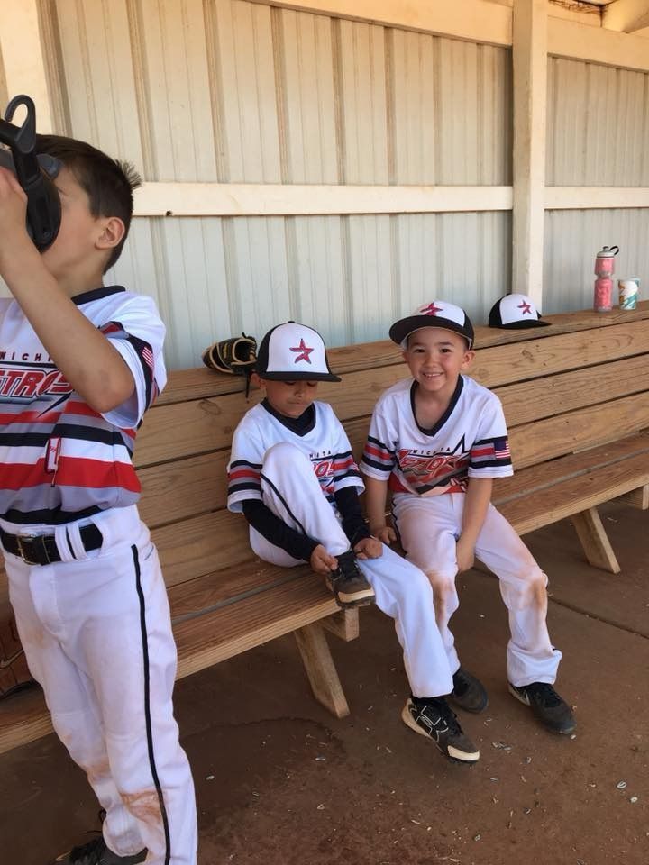 Three children in white baseball uniforms sit and stand by a wooden bench in a dugout.