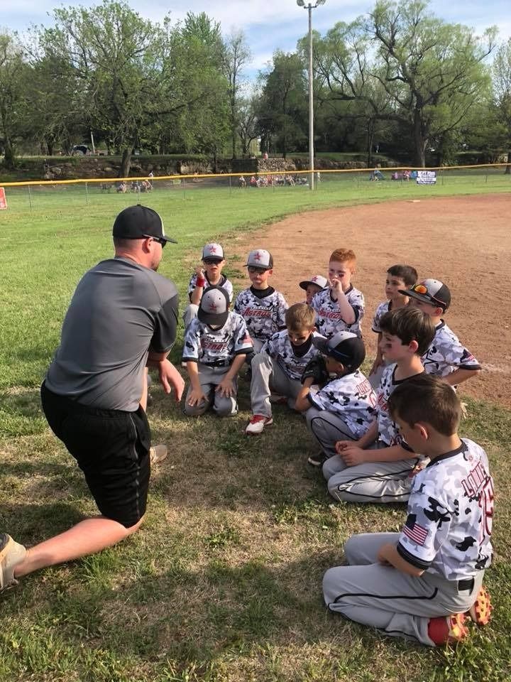 Coach kneels before a youth baseball team seated in a grassy field during practice.