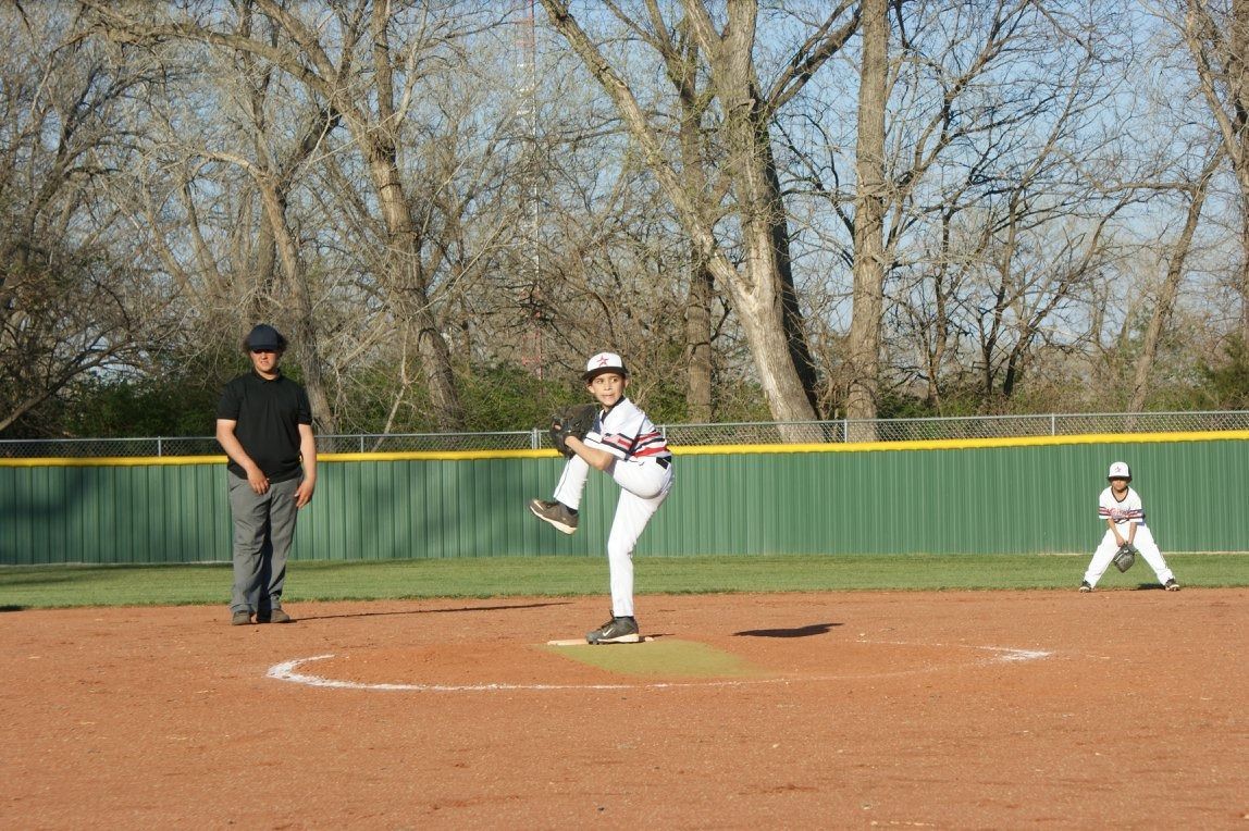 Youth baseball pitcher on mound mid-throw, with umpire and runner in a field under bare trees.
