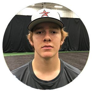 Young baseball player in a white cap and dark shirt standing indoors against black netting.