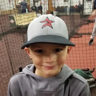 Smiling child in a gray hoodie and white cap with red star, standing indoors by a netted sports area
