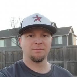 Man in a gray cap with a red star, standing in front of a wooden fence and houses outdoors.