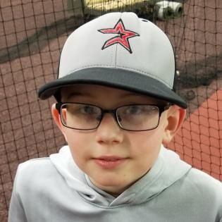 Child wearing glasses and a white cap with a red star, standing in front of a netted sports field.