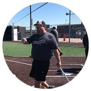 Coach in black shirt and cap walking on a baseball field, holding a bat near home plate