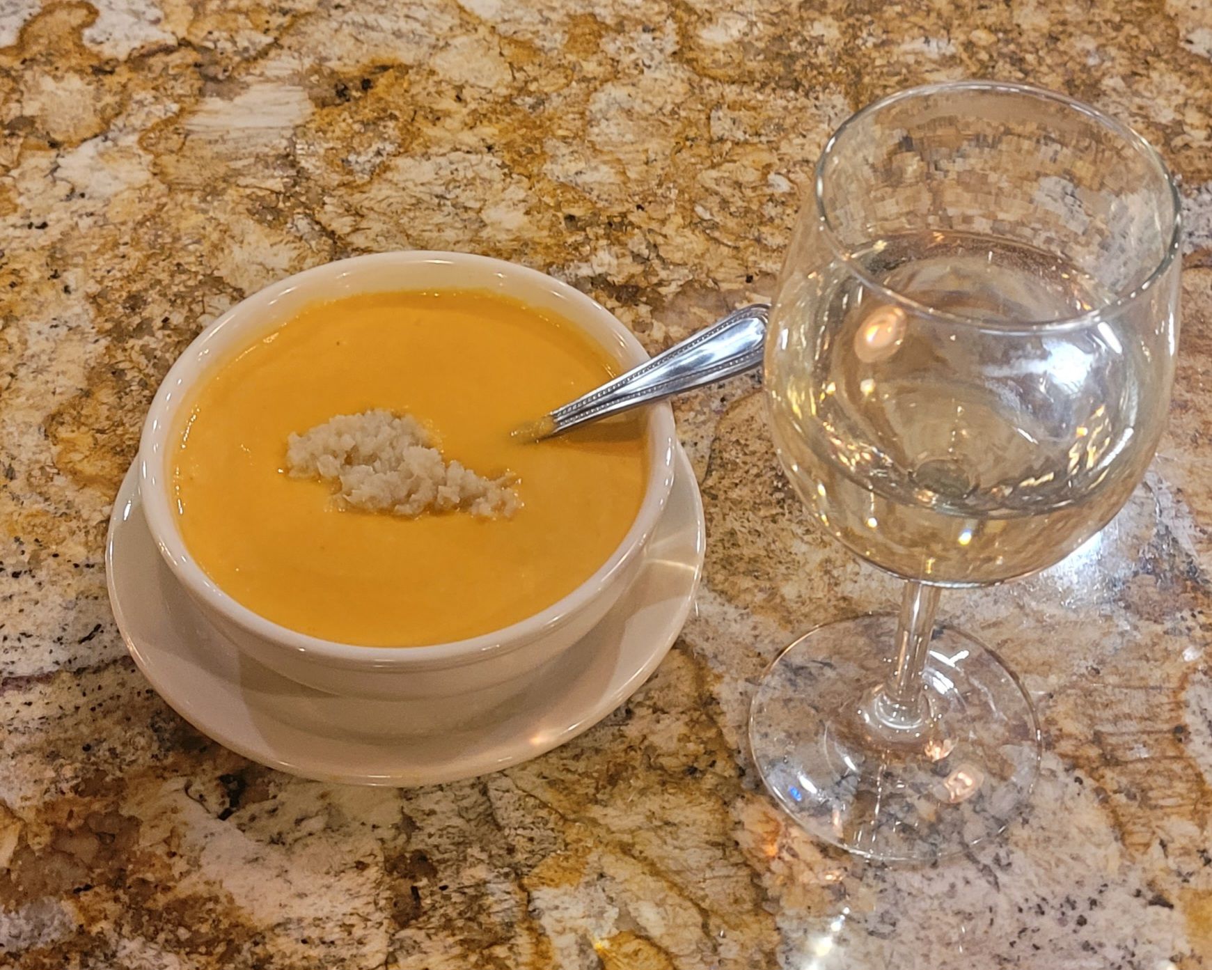 Orange soup in a white bowl with a spoon beside a glass of water on a granite countertop