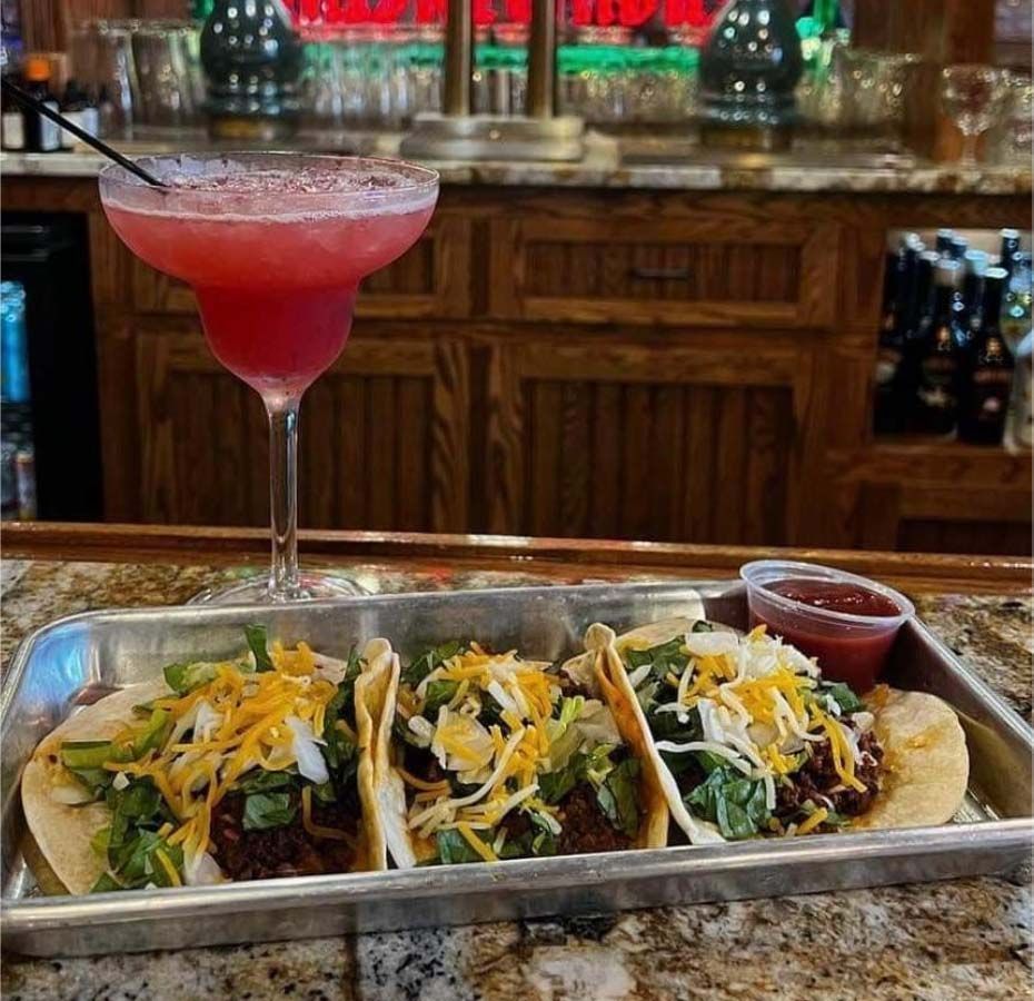 Tray of tacos with toppings beside a pink cocktail at a bar