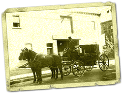 Horse-drawn carriage outside a building. Two horses stand in front of the carriage; a person sits inside.