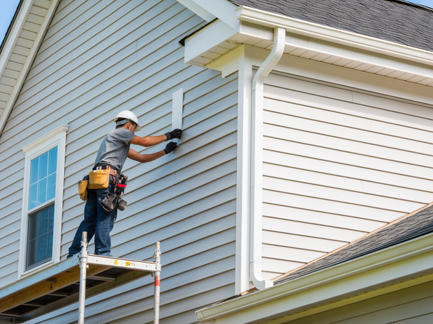 Worker painting house siding on scaffolding beside white and gray exterior walls