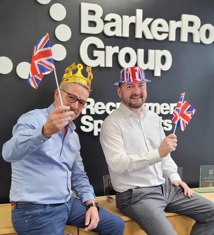 Two men are sitting in front of a barker group sign
