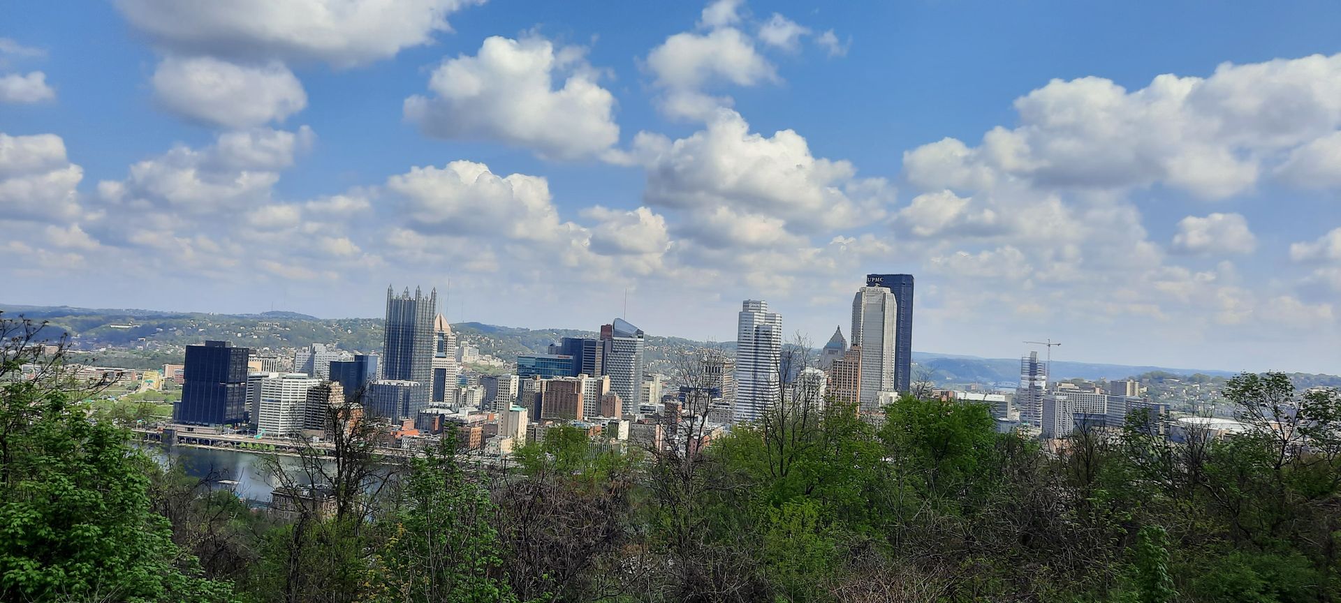 A city skyline with trees in the foreground and a blue sky with clouds in the background.