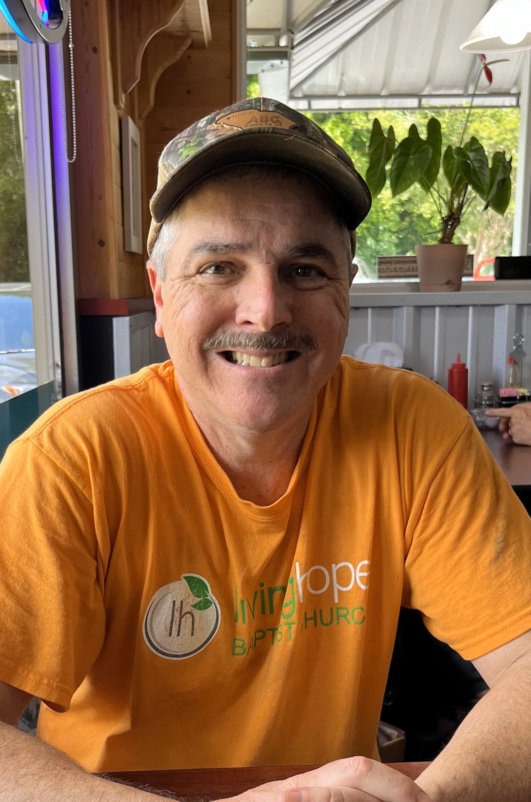A man wearing a hat and an orange shirt is sitting at a table.