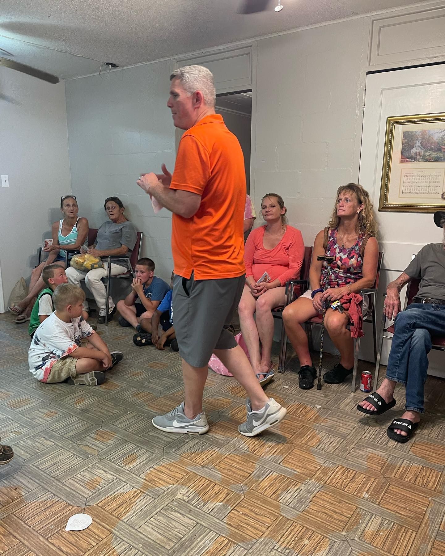 A man in an orange shirt is standing in front of a group of people sitting in chairs.