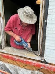 A man in a hat is using a saw to cut a hole in a window.
