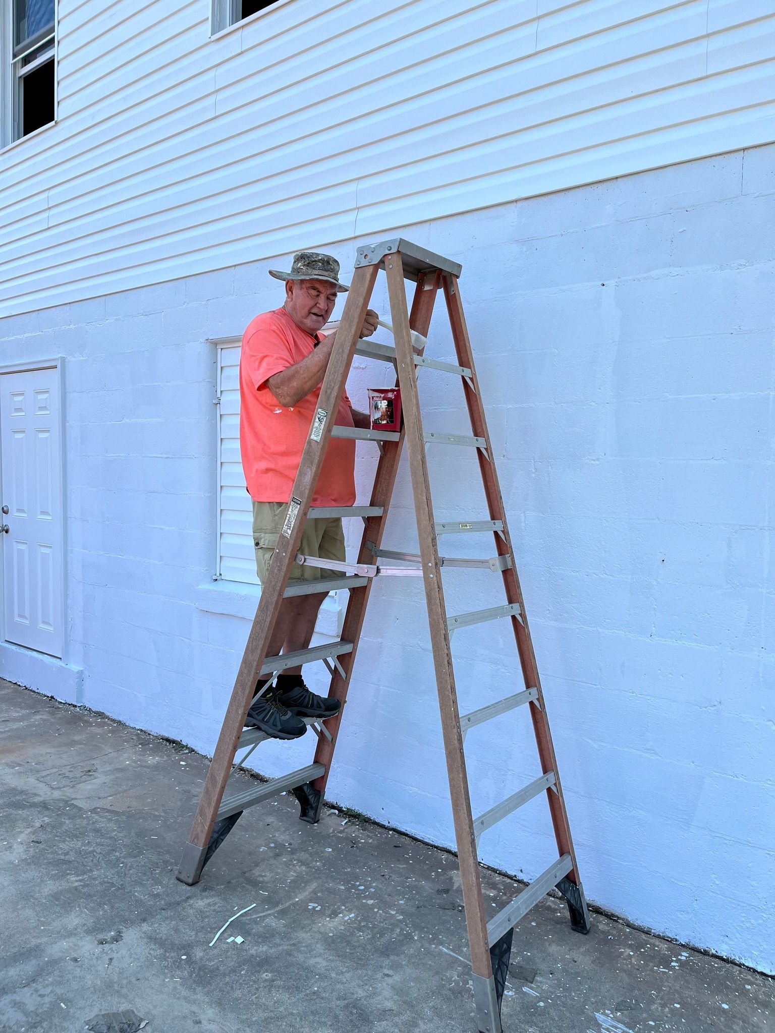 A man is standing on a ladder painting the side of a building.