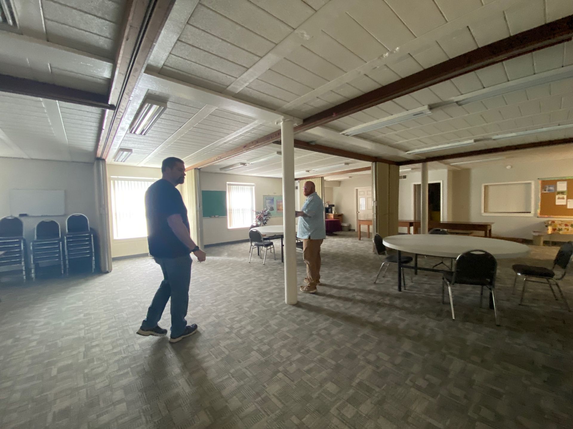 Two men are standing in an empty room with tables and chairs.