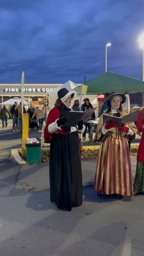 Carolers in period costumes singing carols outside a store under a cloudy sky.
