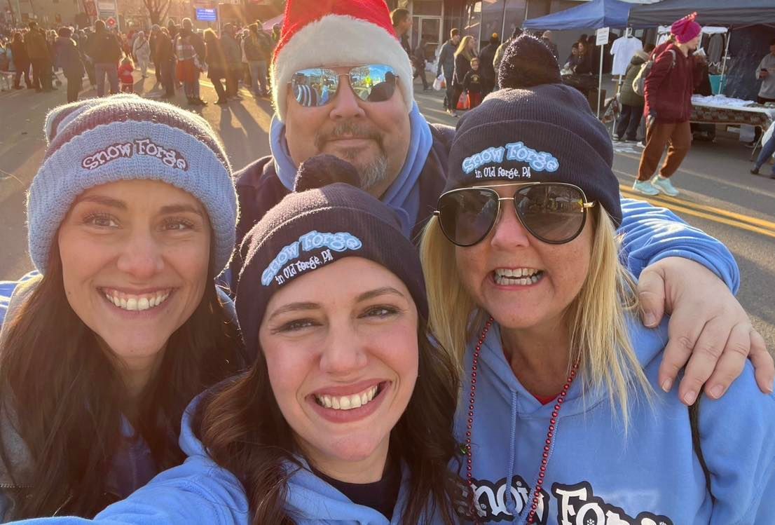 Four people in light blue hoodies and beanies with a logo take a selfie outdoors.