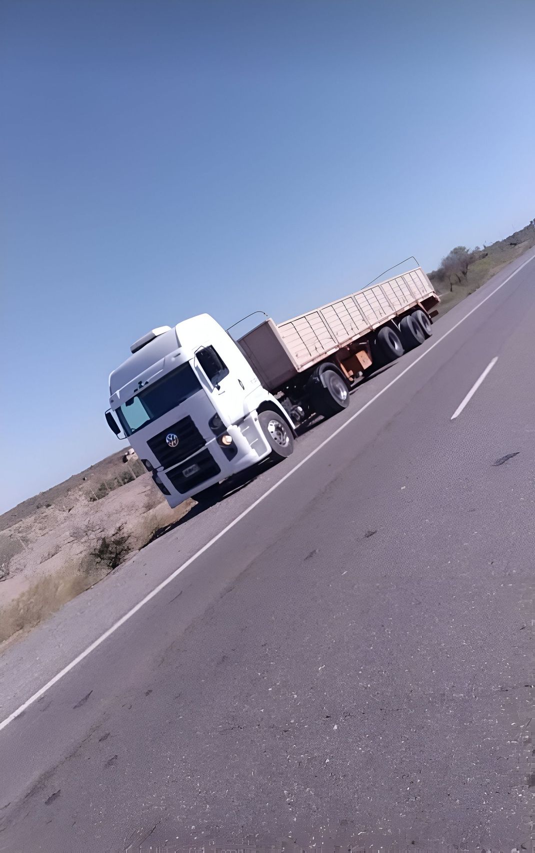 Un camión semirremolque blanco arrastra un remolque marrón por una carretera abierta bajo un cielo azul.
