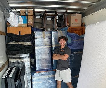 A mover smiles with arms crossed in a packed moving truck, surrounded by furniture and boxes.