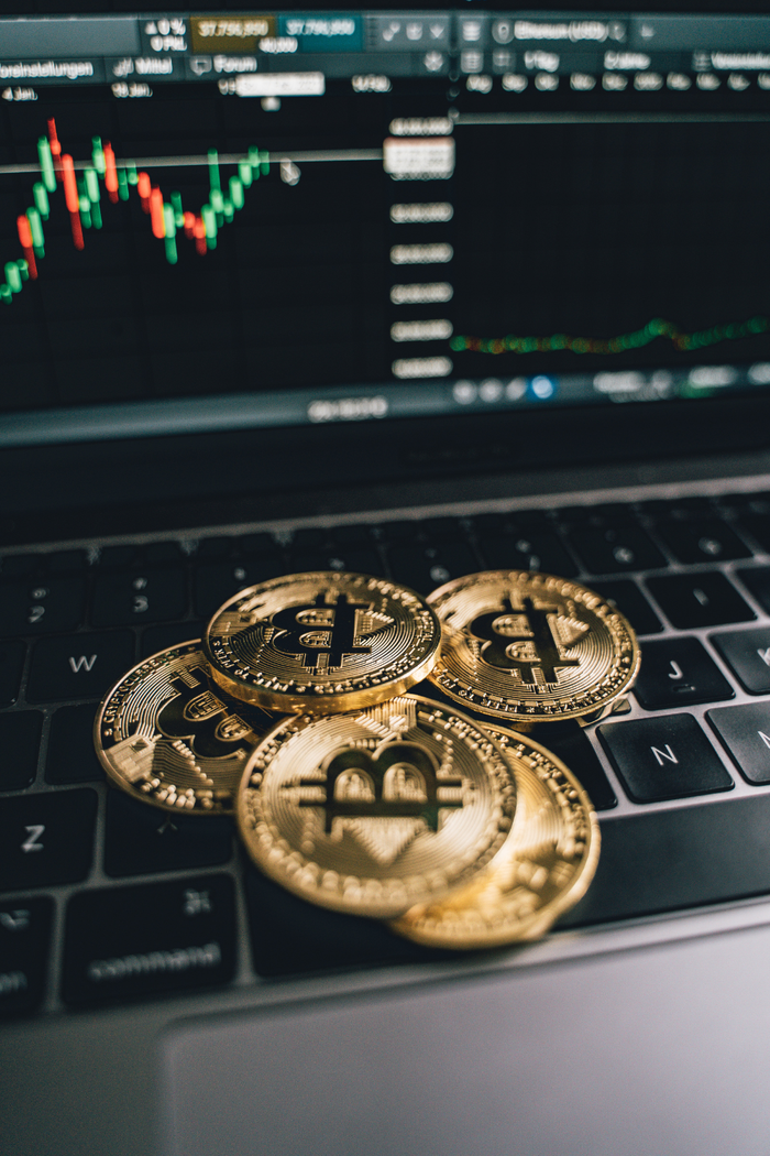 PHYSICAL COINS WITH THE BITCOIN LOGO SITTING ON TOP OF A COMPUTER WITH STOCKS GRAPHS IN THE BACKGROUND