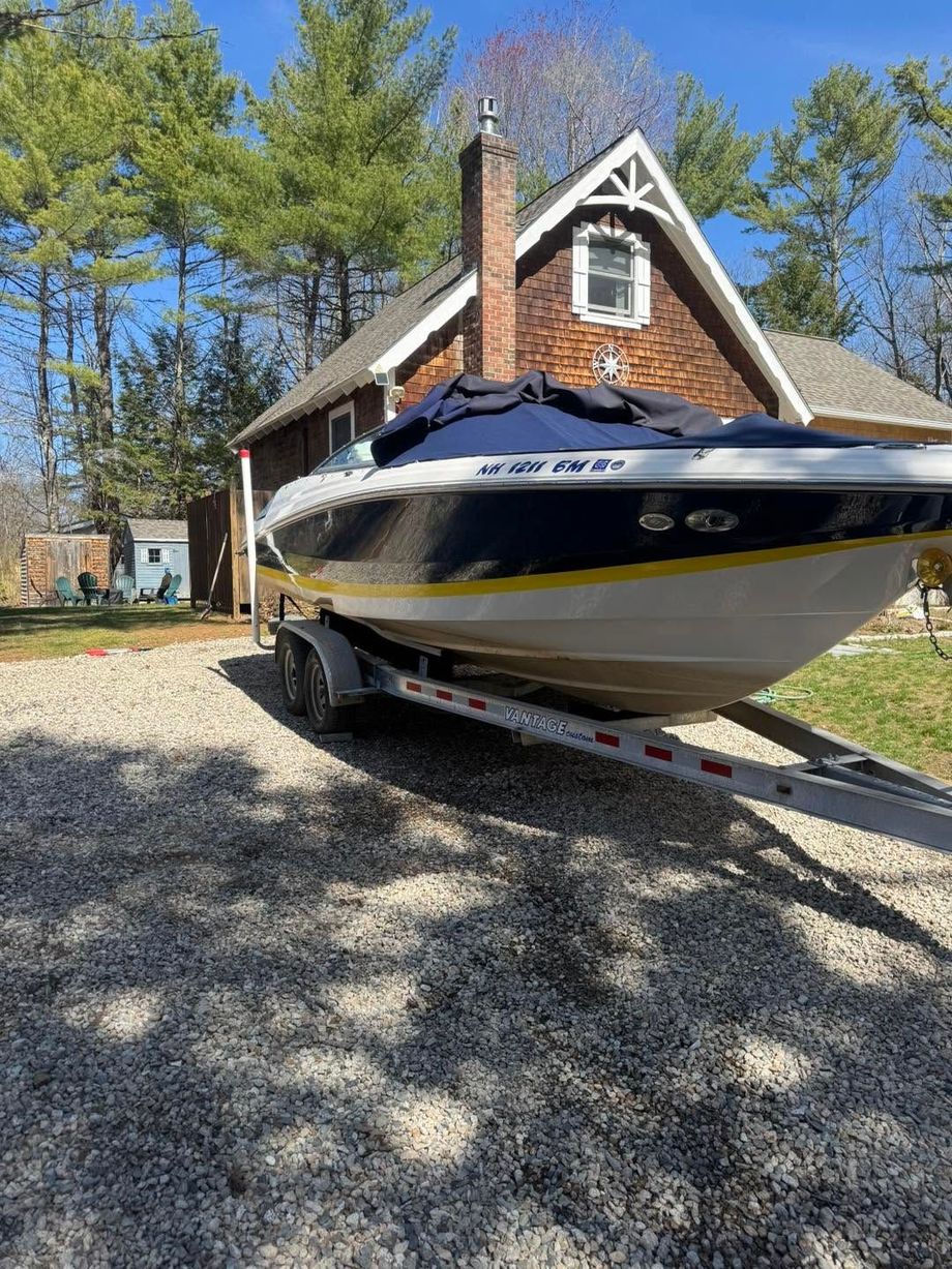 White and navy speedboat on a trailer in a driveway beside a brick house and pine trees