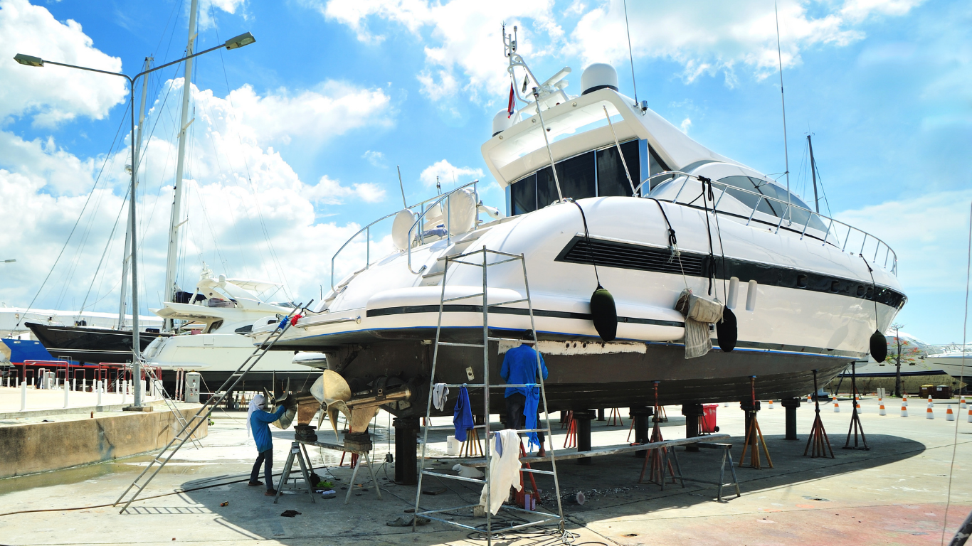 Large white yacht on supports in a dry dock with workers underneath and a bright blue sky