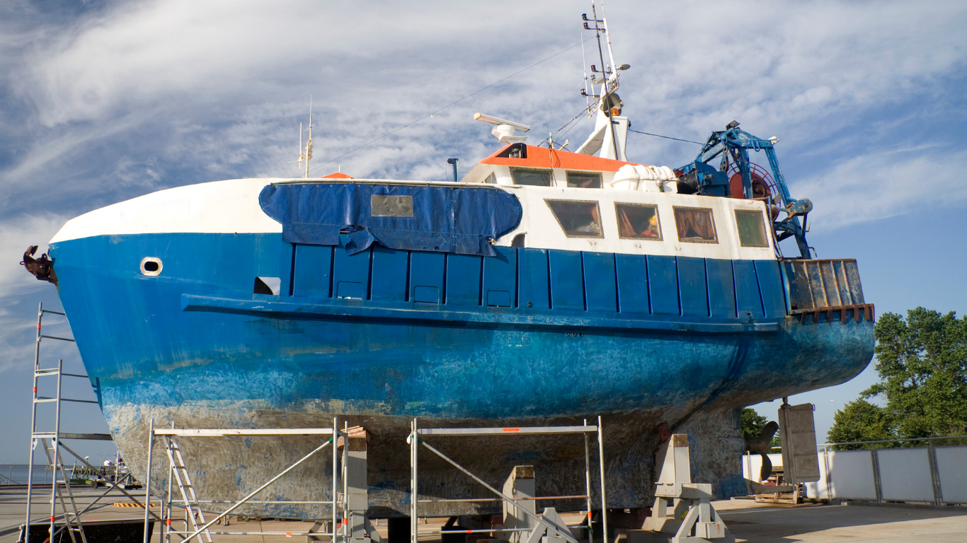 Blue and white boat on supports in a boatyard, with scaffolding and equipment around it