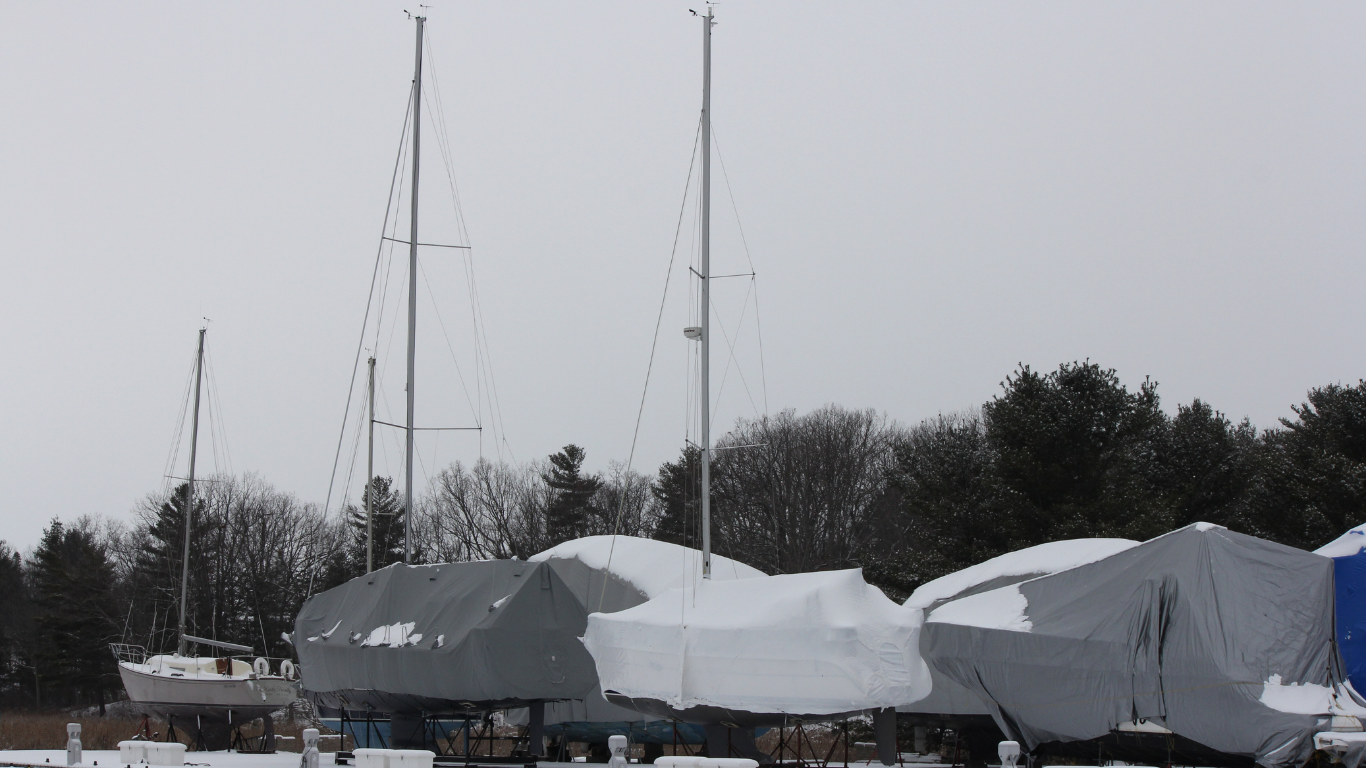 Snow-covered boats stored outdoors with tall masts against trees and a gray sky
