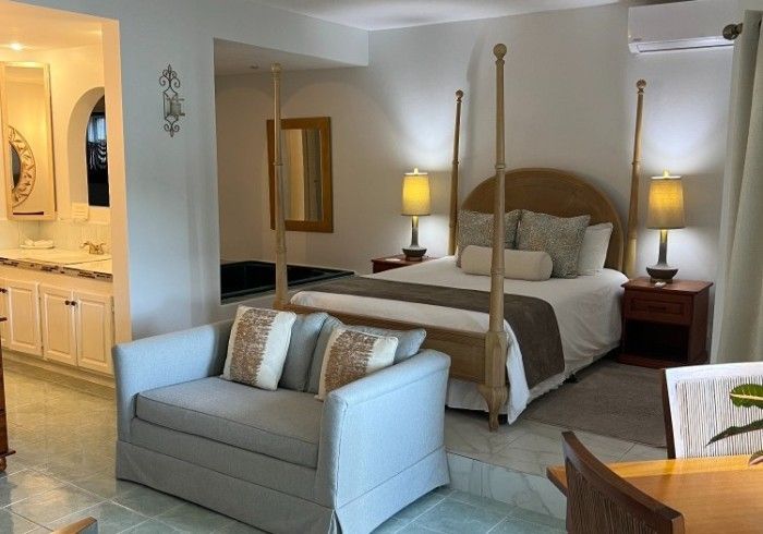 Bright bedroom suite with four-poster bed, gray sofa, bedside lamps, and an adjoining bathroom vanity.