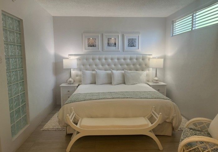 Bright white bedroom with tufted bed, lamps, framed art, and a bench at the foot of the bed.