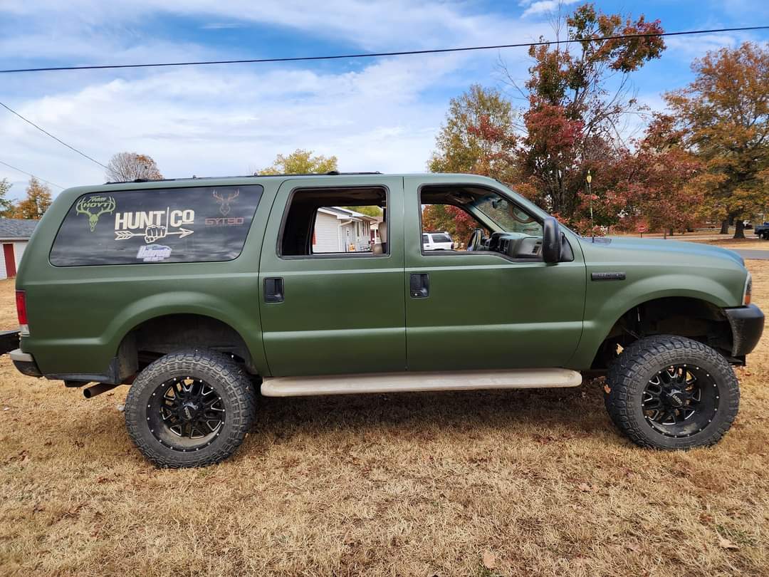 A matte olive green Ford Excursion SUV with black off-road wheels, parked in a dry, grassy field.