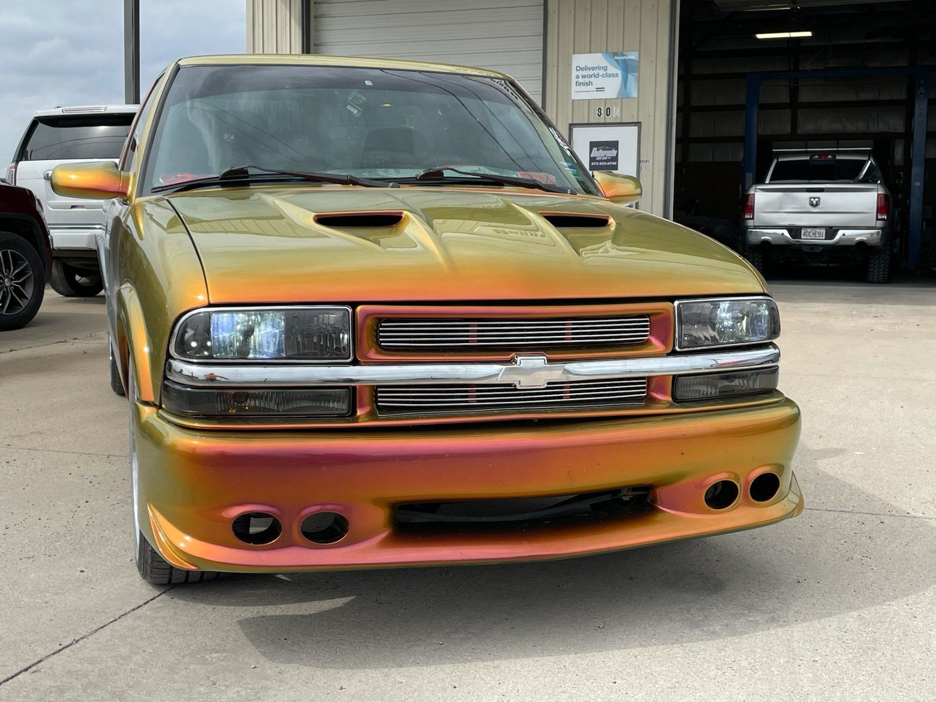 A custom truck with a color-shifting, iridescent gold-to-orange finish, parked in front of a service garage.