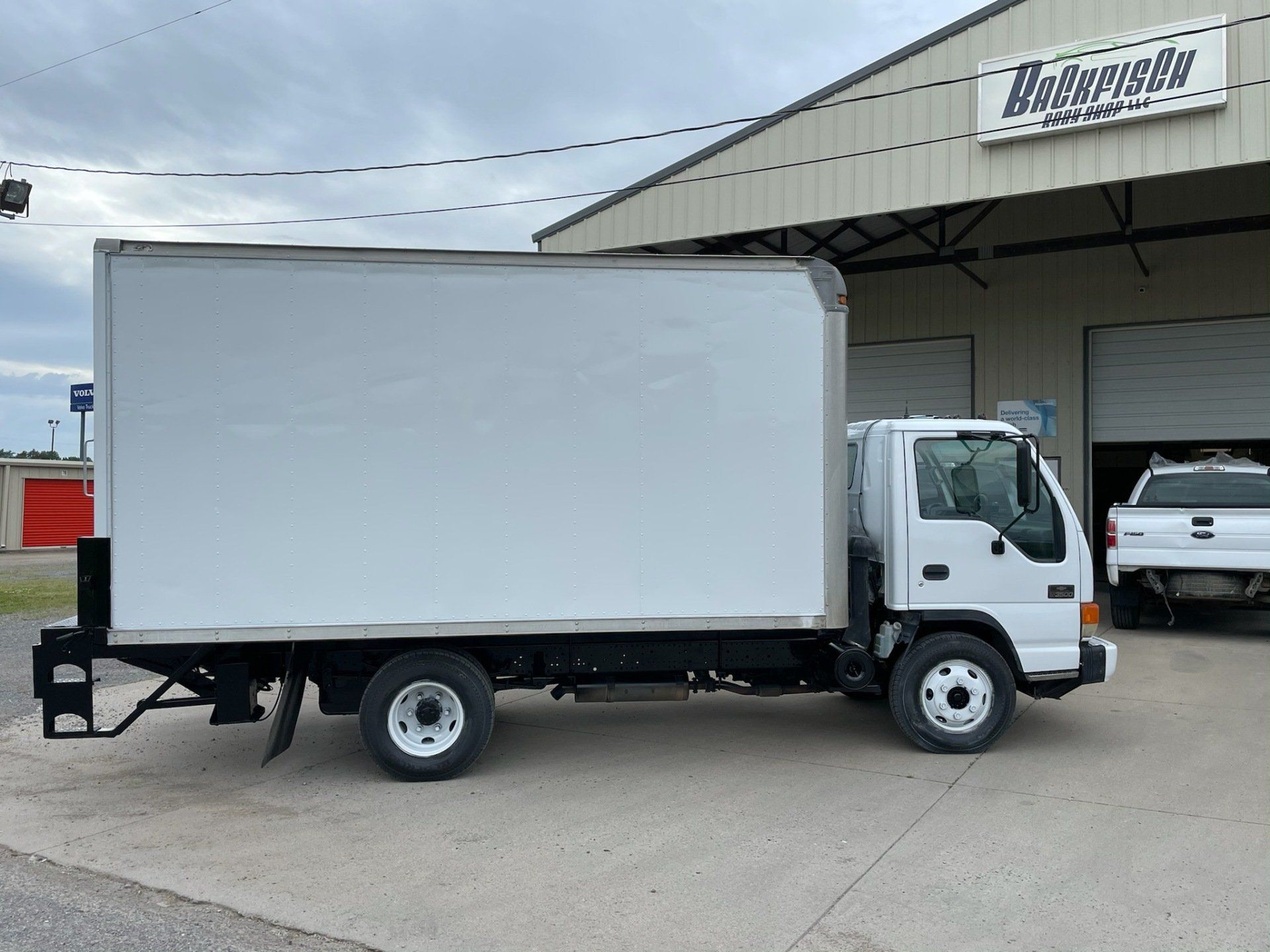A white box truck parked in front of a metal-sided building with another truck partially visible in the background.