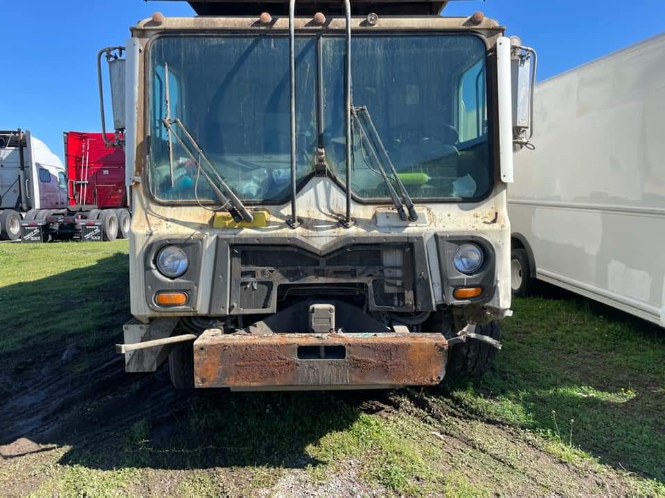 A front view of an old, weathered, light-colored cab-over truck parked on grass under a clear blue sky.