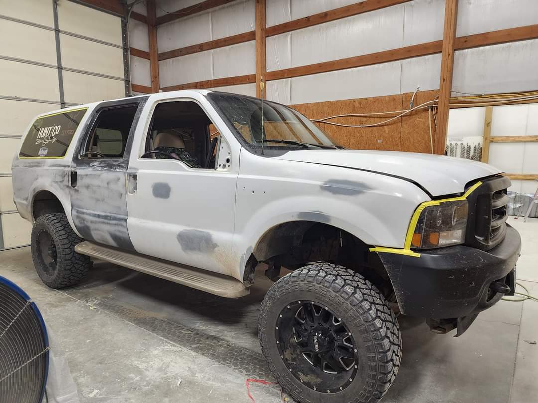 A white Ford Excursion undergoing body work in a garage, featuring grey primer spots and large black off-road tires.