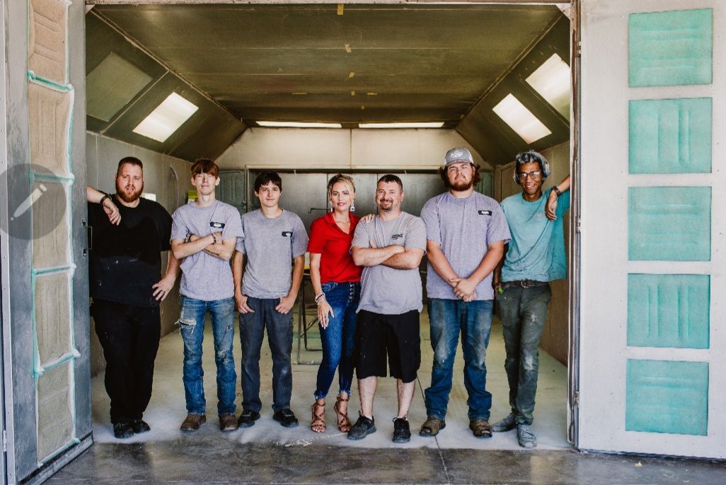 Seven people stand in a line inside a brightly lit, industrial paint booth with metallic walls and green filters.