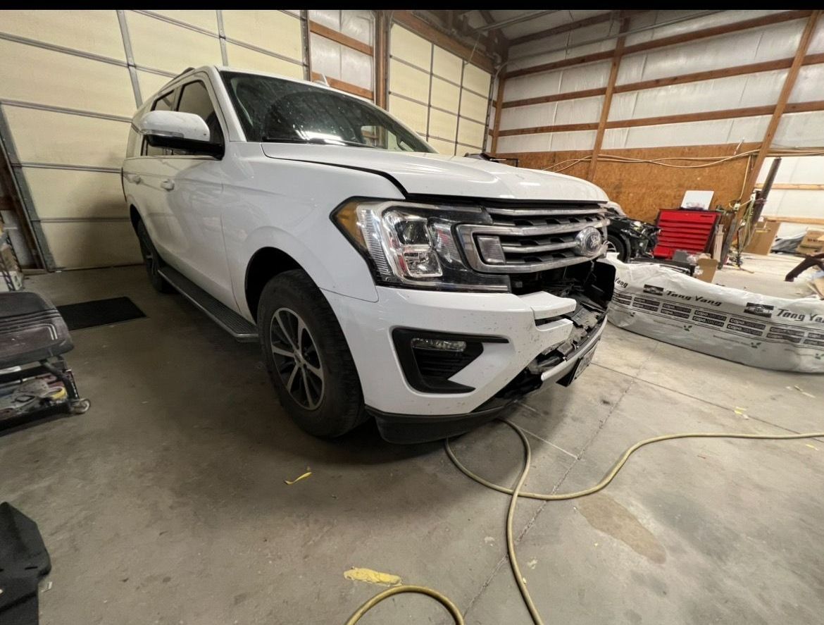 A white SUV parked in a workshop with a damaged front bumper and grille.