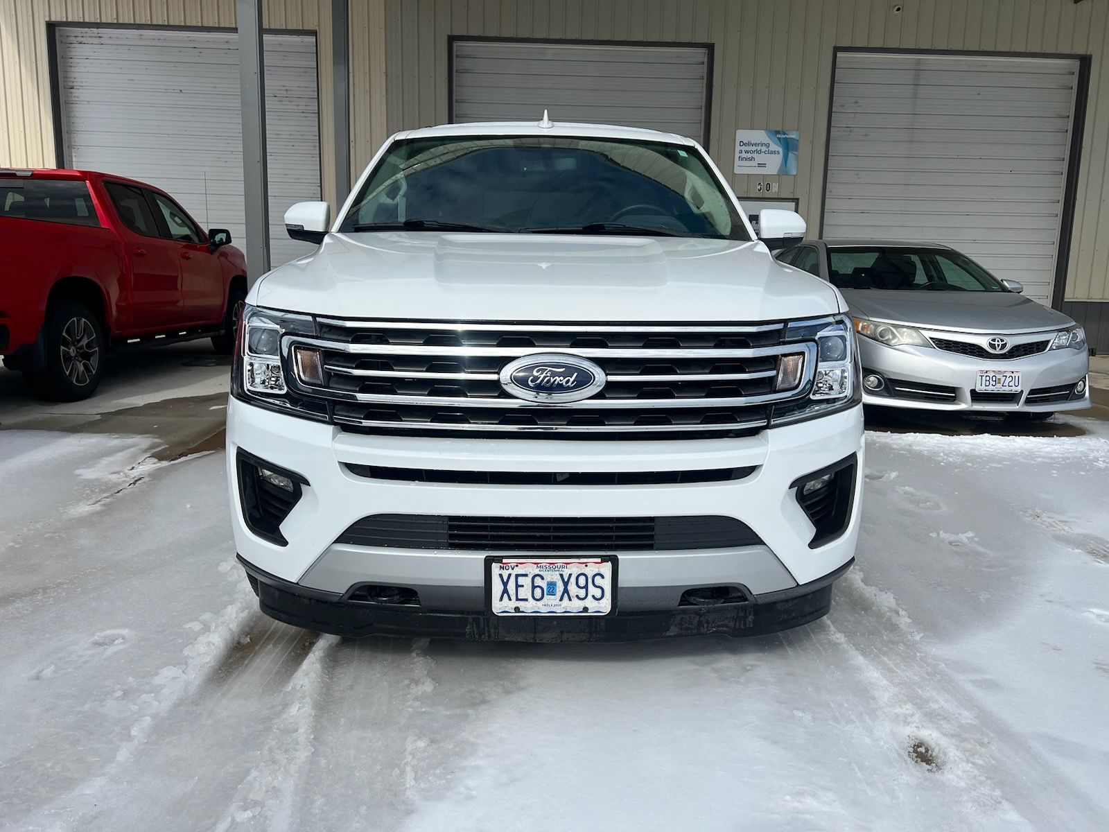 A white Ford SUV parked in a snowy lot, flanked by a red pickup truck and a silver sedan.
