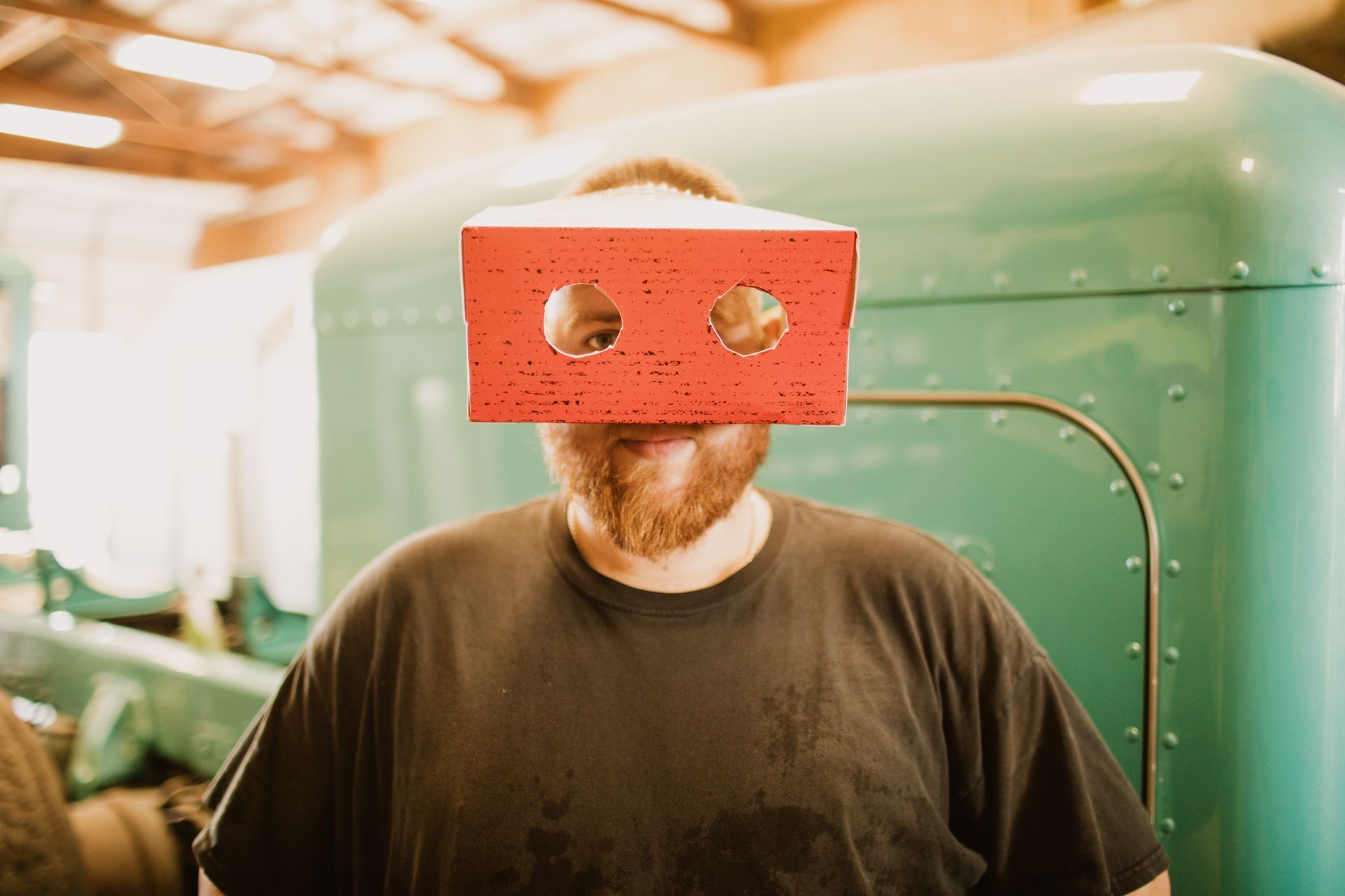 A person in a black t-shirt wears a red brick with two holes cut out for eyes as a mask, standing before a green truck.