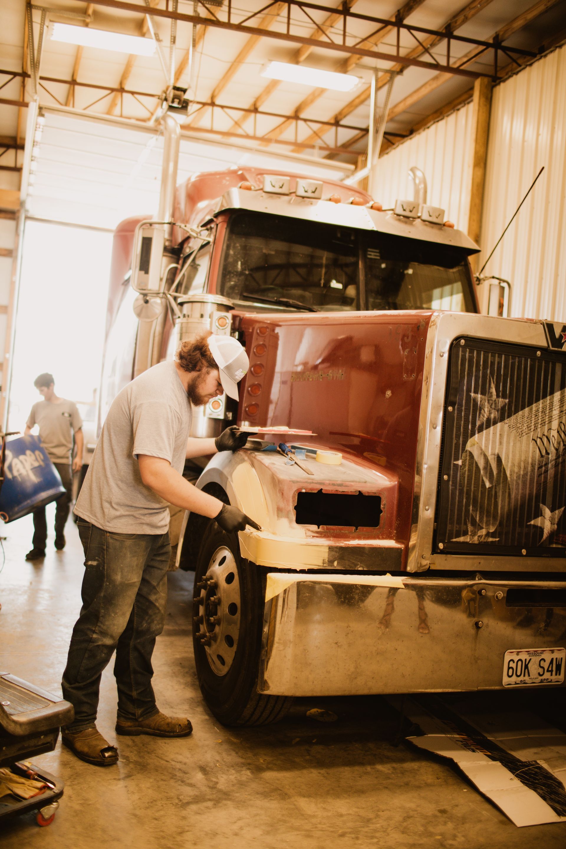 A person wearing a hat and gloves works on the front corner of a maroon semi-truck inside a garage.