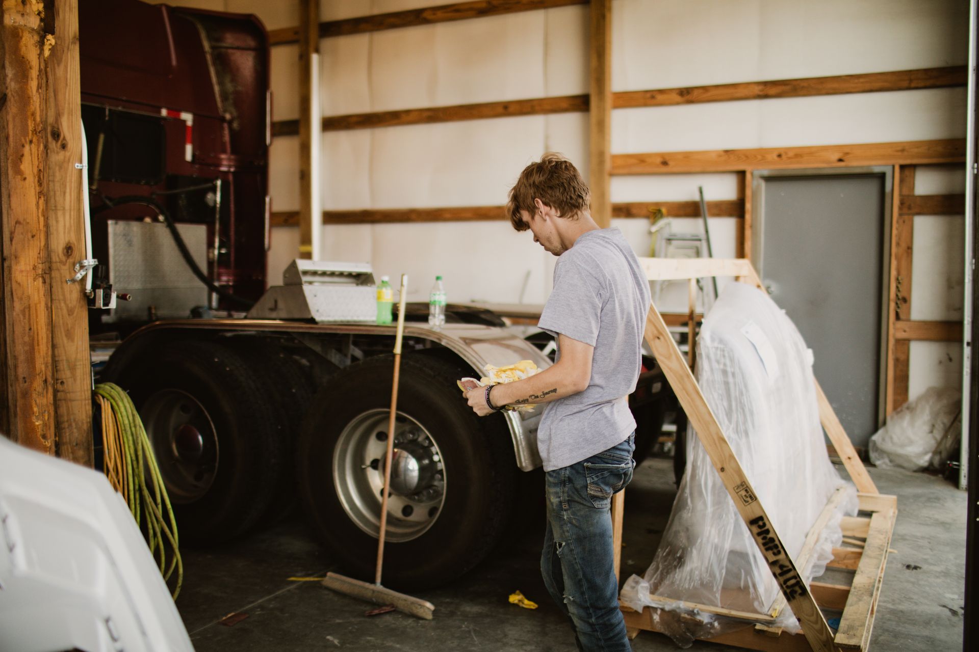 A person in a light shirt and jeans works on a truck's wheel well inside a large, wooden-framed workshop.