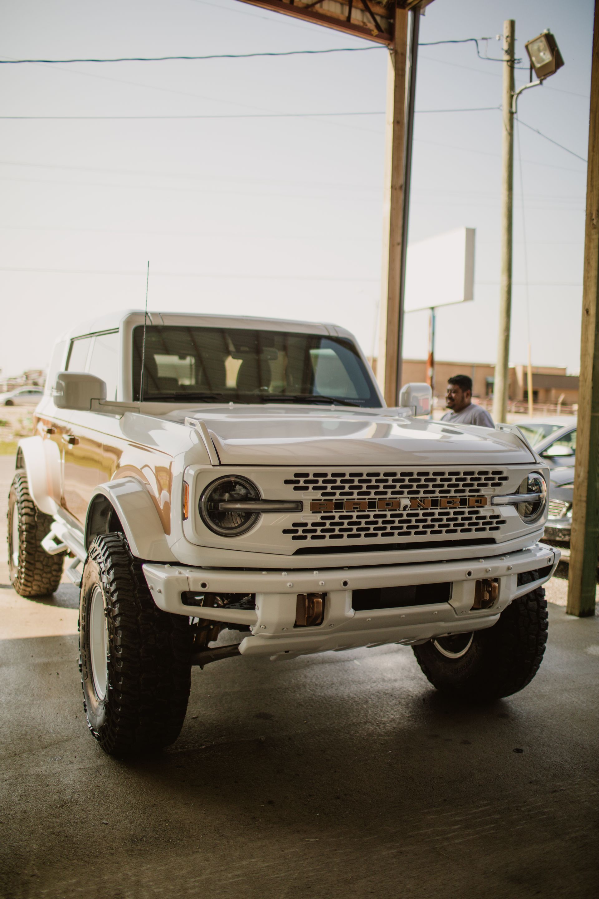 A modified white Ford Bronco with oversized tires parked under a shelter in bright daylight.