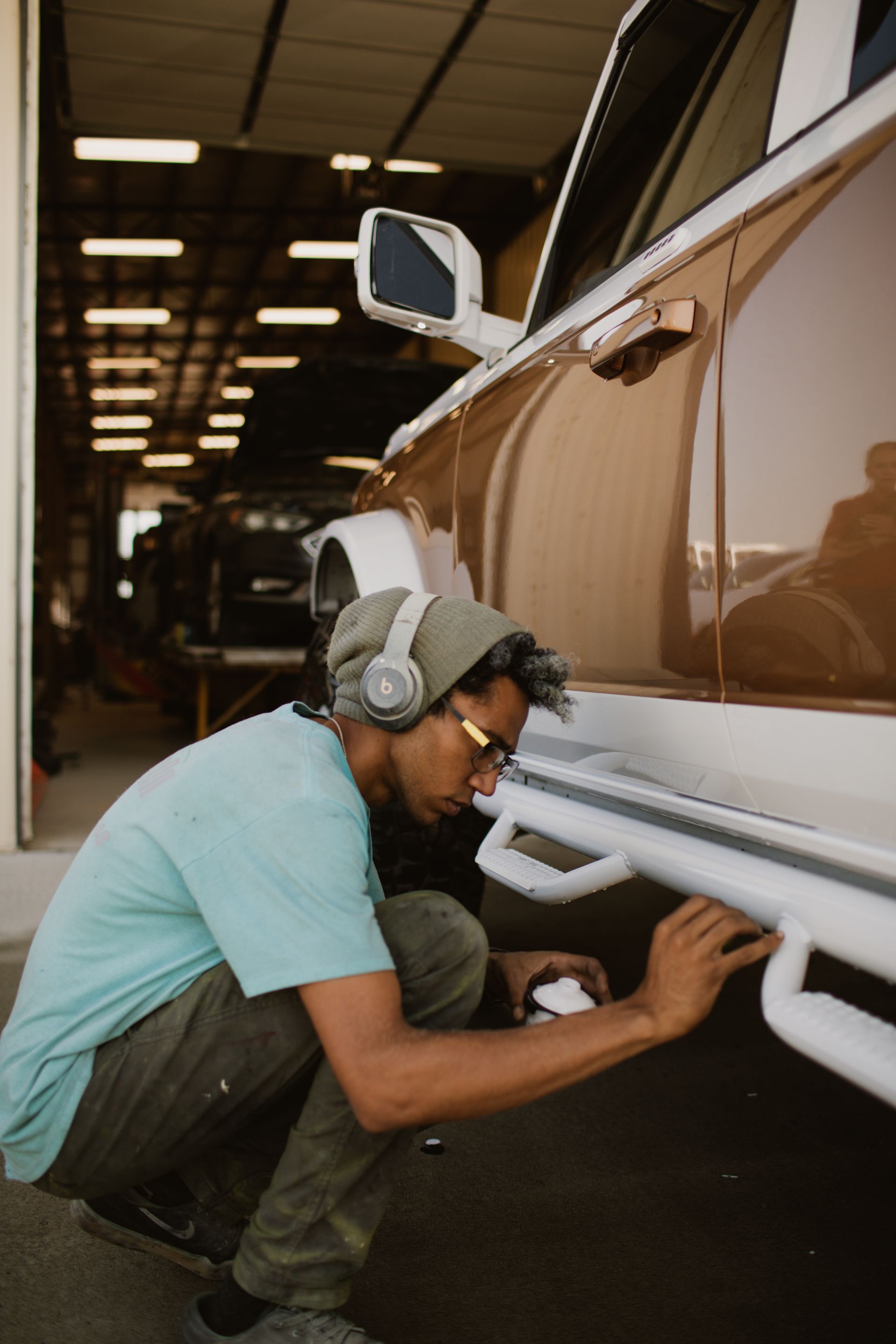 A person in a beanie and blue t-shirt kneeling to paint the white running board of a bronze-colored vehicle in a garage.