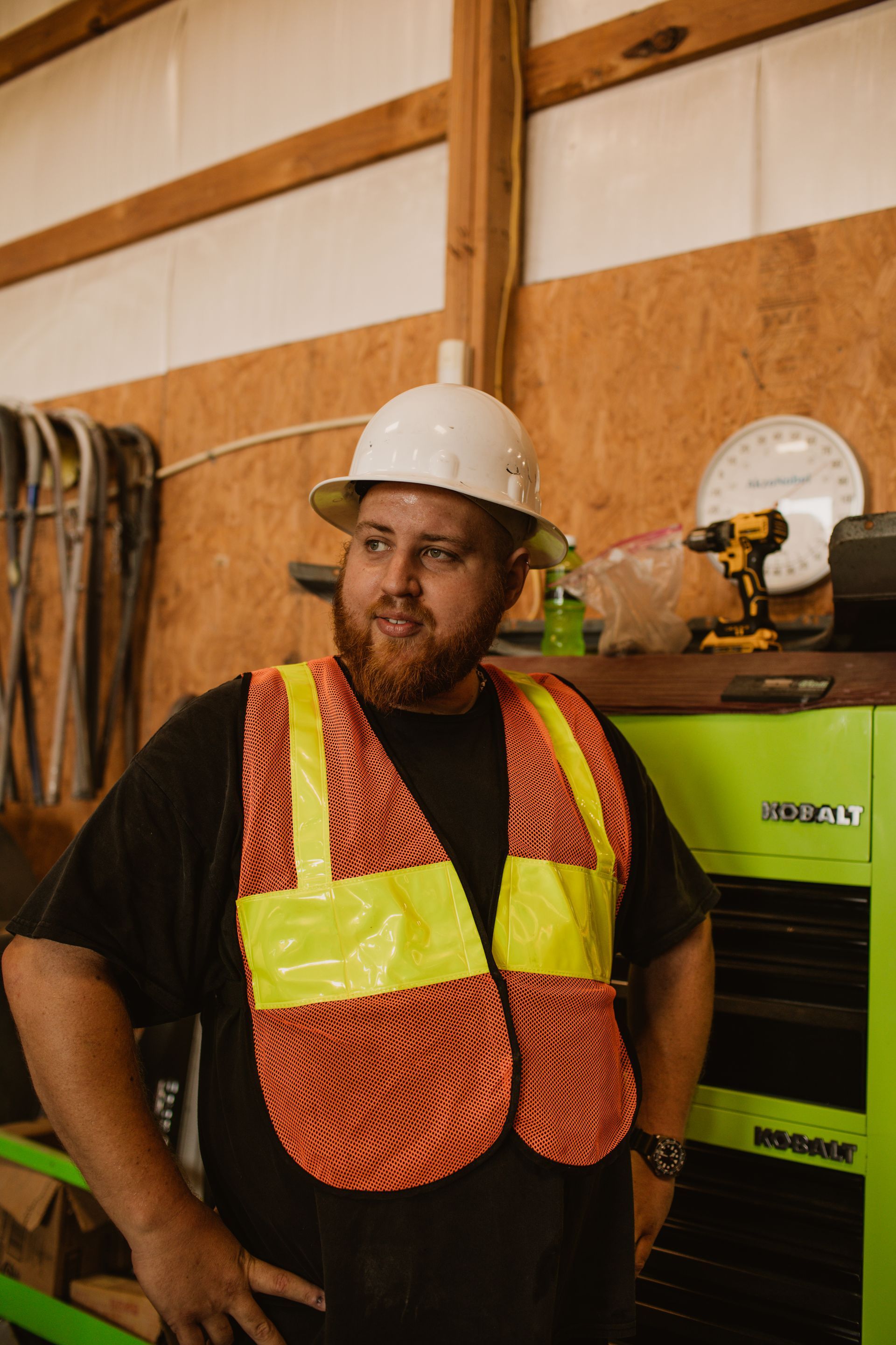 A person with a beard, wearing a white hard hat and an orange high-visibility vest, stands in a workshop.