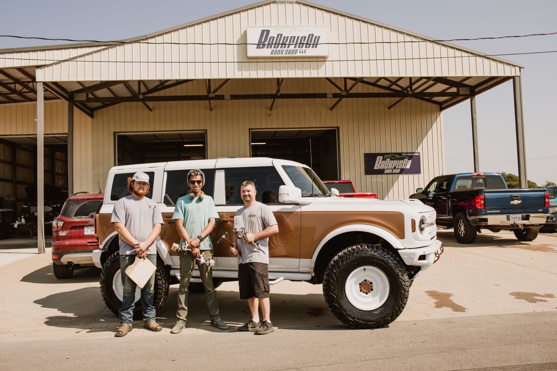 Three people stand before a customized white and tan Ford Bronco parked outside a mechanic shop.