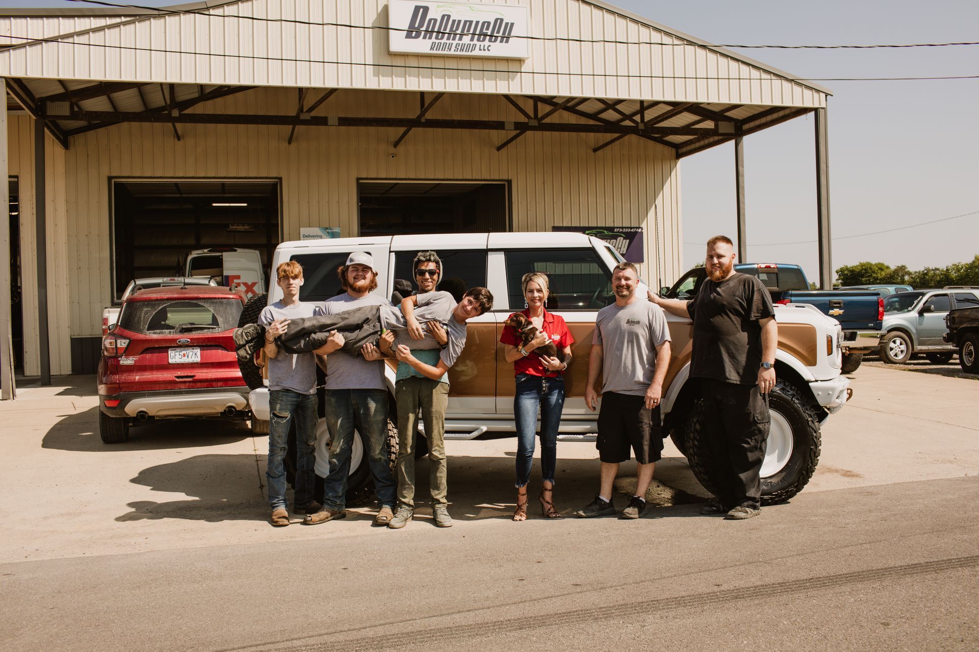 Six people pose in front of a white SUV with wood-paneled sides outside a garage labeled 