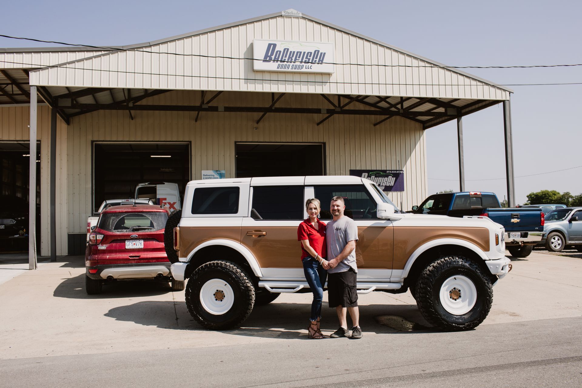 A couple stands in front of a modified white and gold Ford Bronco at an auto shop.